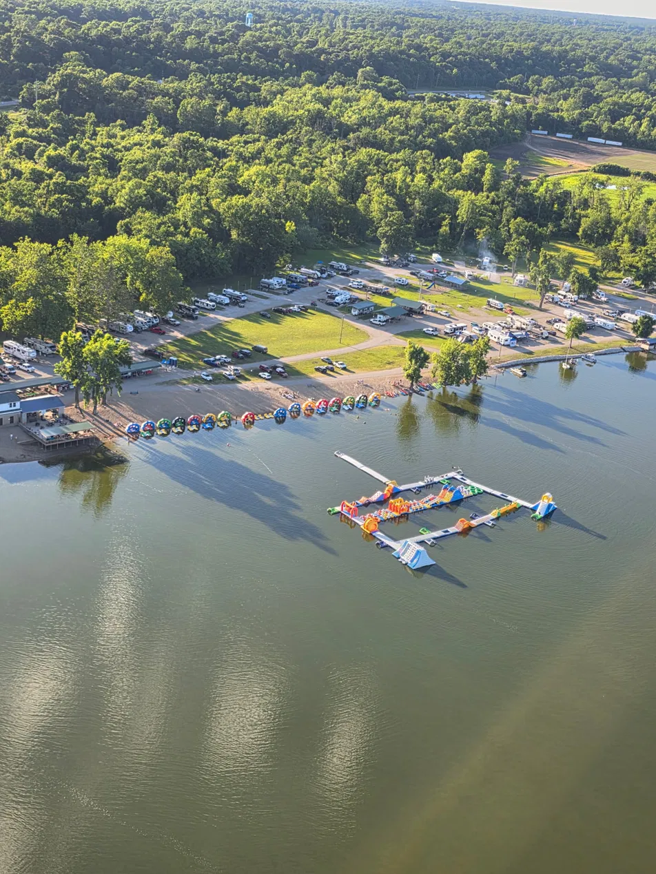 Aerial view of Lazy L Lake Campground showing RV sites, the sandy swimming beach lined with kayaks and floats, and the inflatable aqua park on the 75-acre lake